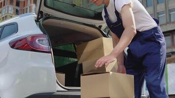 Moving and delivery of manufactured goods. The assistant puts the cargo in the craft boxes in the trunk of the car and closes the car door. Side view video