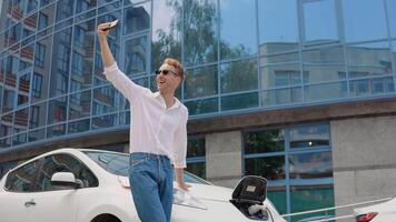 Stylish modern young curly man stands near an electric car charging and takes a selfie with his smartphone video