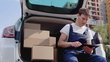 The courier sits on the edge of the open trunk, filled with boxes of goods and fills out documents video