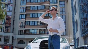 Stylish modern young curly man in jeans and a white shirt stands near an electric car with a charger in hand in the yard of a residential complex video