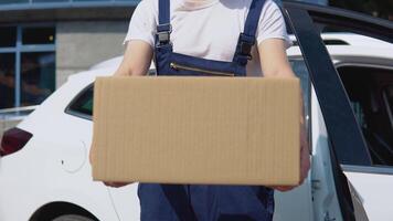 A delivery worker in a white T-shirt and blue jumpsuit stands by an open car and gives a box to recipient in the direction to the camera video