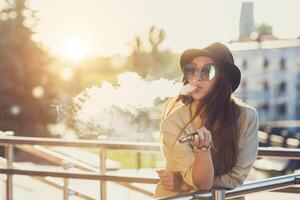 bonito joven hipster mujer en negro sombrero vape cigarrillo electrónico, vaping dispositivo a el puesta de sol. tonificado imagen. foto