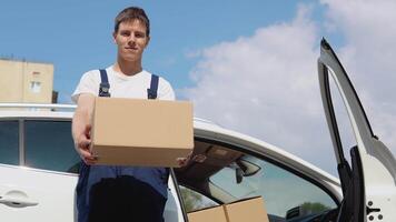 en mouvement et livraison de fabriqué marchandises. le chauffeur remplit dans le cargaison les documents et regards dans le caméra. un employé détient une boîte dans le sien mains et des stands suivant à une voiture rempli avec colis video