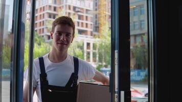 A courier in a white T-shirt and blue jumpsuit holds a box in one hand, and with the other opens the glass door of a modern building and goes inside. Delivery of goods to the office video