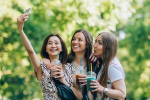 Happy friends in the park on a sunny day . Summer lifestyle portrait of three multiracial women enjoy nice day, holding glasses of milkshakes. Taking picture on mobile phone. Best friends girls having photo