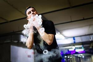 Man in cap smoke an electronic cigarette and releases clouds of vapor performing various kind of vaping tricks photo