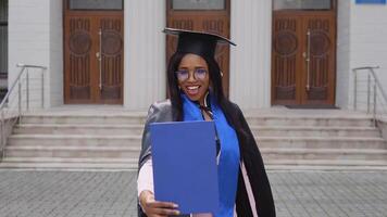 un tímido africano americano hembra graduado en un negro manto de un maestría sombrero poses para el cámara. un importante evento. joven especialista video