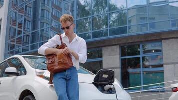 Stylish modern young curly man with a stylish city briefcase walks away from his electric car, leaving the car to charge video