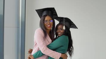 Two girlfriends graduates in festive costumes and master's hats hug and look at the camera with a smile video