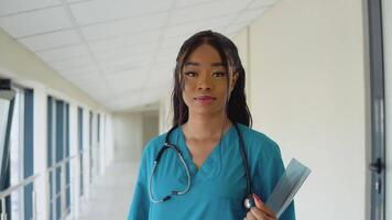 uma jovem africano americano médico mulher dentro uma azul terno carrinhos dentro a corredor do a clínica e parece para dentro a Câmera. a médico é segurando a raio X video