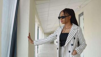 Glamorous African American businesswoman in black glasses and white striped suit standing by the window in the hallway of the office center video