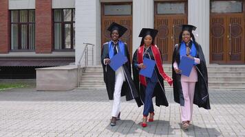 Happy graduates of a university or college of African American nationality with blue diplomas in their hands walk forward to the camera from the entrance to the university video