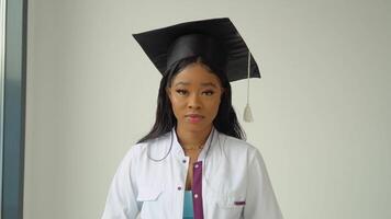 un joven africano americano hembra graduado en un maestría sombrero y blanco médico vestido poses para el cámara. un importante evento. joven especialista video