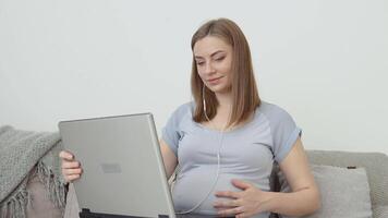 uma grávida mulher dentro casa roupas é sentado em uma sofá e usando uma computador portátil. grávida mulher trabalhador autonomo. terceiro trimestre do gravidez. preparação para parto e Cuidado do a recém-nascido bebê video