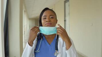 Jeune africain américain médecin femme dans bleu costume et blanc manteau supprime jetable stérile visage masque video