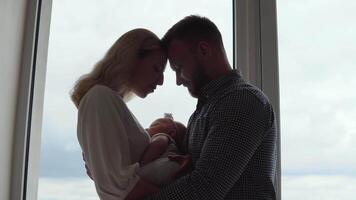 The mother and father stand in front of the window facing each other and hug while the child sleeps between them in the mother's arms. Fatherhood and motherhood. Traditional family video