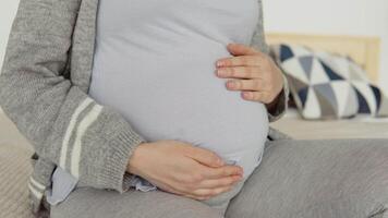 Close up of pregnant woman in home clothes sitting on a double bed. Dried flowers in the interior. The pregnant woman is stroking her belly. Third trimester of pregnancy video