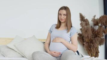 zwanger vrouw in huis kleren zittend Aan een dubbele bed. droog bloemen in de interieur. de zwanger vrouw is strelen haar buik. derde trimester van zwangerschap video