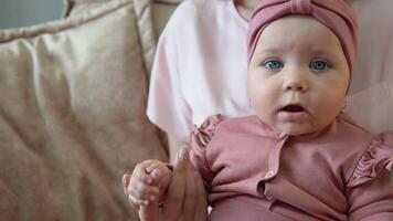 Baby girl with blue eyes in a cute pink suit sits in mom's lap. Mother and baby in pink clothes sitting on a soft light sofa facing the camera video