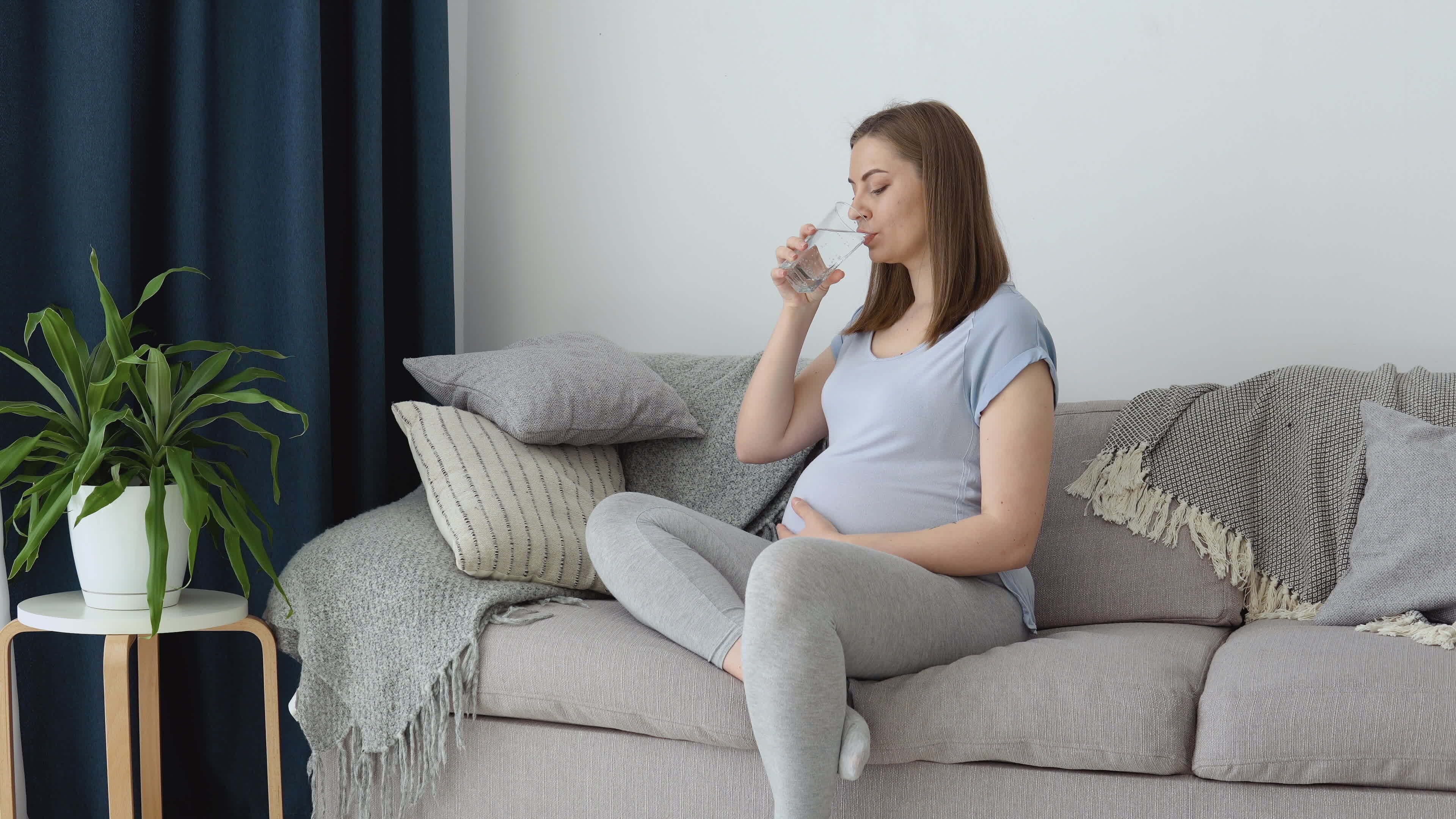 A pregnant woman in home clothes sits on the couch and drinks clean