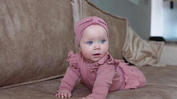 A baby girl with blond hair and blue eyes in pink clothes lies on her tummy and swings in different directions. Baby on a light beige sofa video