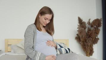 embarazada mujer en hogar ropa sentado en un doble cama. seco flores en el interior. el embarazada mujer es acariciando su barriga. tercero trimestre de el embarazo video
