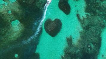 Aerial top view captures heart-shaped coral reef embraced by vibrant turquoise waters in tropical paradise located in Caribbean Sea. Boat carrying tourists sails past. Zoom out video