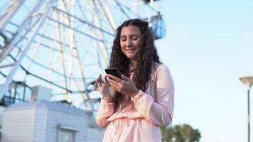 un hermosa joven niña con oscuro oscuro pelo usos un teléfono inteligente en pie cerca un grande ferris rueda en un diversión parque. lento movimiento video