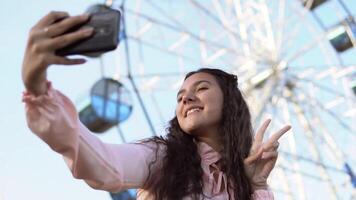 A beautiful girl with long hair in a dress makes selfie using a phone while standing near the Ferris wheel. slow motion. Portrait video