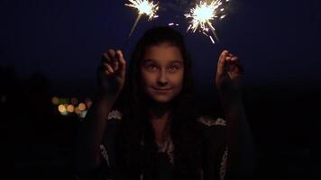 joven hermosa niña con largo oscuro pelo sostiene fuegos artificiales a noche en el antecedentes de el ciudad y se regocija lento movimiento. video
