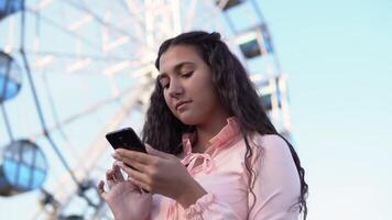 uma lindo jovem menina usa uma Smartphone em pé perto uma ampla ferris roda dentro a diversão parque. lento movimento video