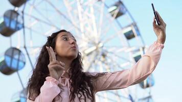 A beautiful girl with long hair in a dress makes selfie using a phone while standing near the Ferris wheel. slow motion. video