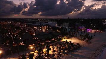 turist hotell mot bakgrund av lila solnedgång upplyst förbi värma ljus av lyktor, på tropisk kust. strand paraplyer och Sol solstolar. Drönare är flygande framåt- och nedåtgående. video
