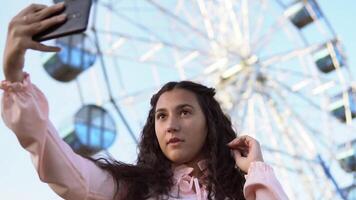 ein schön Mädchen mit lange Haar tut Selfie mit ein Telefon während Stehen in der Nähe von das Ferris Rad. schleppend Bewegung. Porträt video