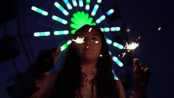 uma lindo jovem menina com grandes Sombrio cabelo carrinhos contra a fundo do uma cidade às noite e detém fogos de artifício dentro dela mãos. lento movimento. hd video