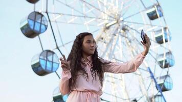 jung schön Mädchen mit lange Haar im ein Rosa lange Kleid macht Selfie mit ein Telefon während Stehen in der Nähe von das Ferris Rad. schleppend Bewegung. video