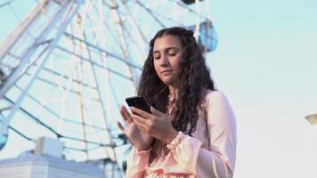 ein schön jung Mädchen mit gegessen Haar Verwendet ein Smartphone Stehen in der Nähe von ein groß Ferris Rad im ein Amüsement Park. schleppend Bewegung video