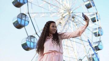 Young beautiful girl with long hair in a pink long dress makes selfie using a smartphone standing near the Ferris wheel. slow motion. video