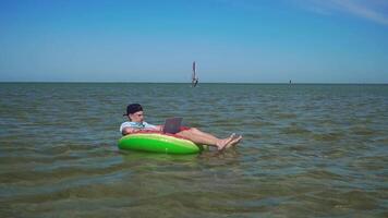 A young business person is enthusiastically working behind a laptop swaying on the waves of sea sitting on a inflatable circle. 4K video