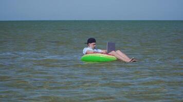 A young business person is enthusiastically working behind a laptop swaying on the waves of sea sitting on a inflatable circle. 4K video