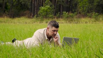 Man Works behind a Laptop lying on the Grass At The Sunny Day. Freelancer Remote Work Concept. 4K video