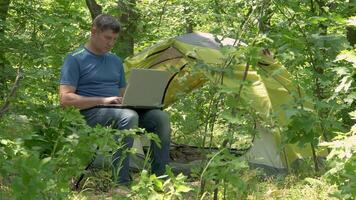 Mens werken achter een laptop in de buurt in de buurt de tent in de Woud. freelancer afgelegen werk concept. 4k video