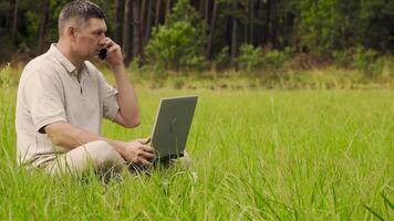 uma homem com uma computador portátil é falando em a telefone em a verde gramado às a ensolarado dia. trabalhador autonomo controlo remoto trabalhos conceito. 4k video