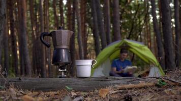 Aromatic coffee is prepared in a geyser coffee maker. A man reads a book in a tent in the forest in the background. Travel and healthy lifestyle concept video