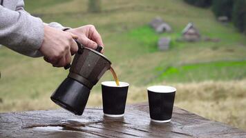 The tourist's hand pours aromatic coffee into a mug from a geyser coffee maker. Wooden houses of shepherds in the background. Travel concept video