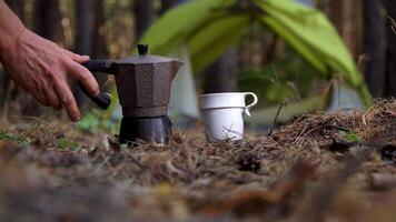 The tourist's hand pours aromatic coffee into a mug from a geyser coffee maker. A tent in the forest in the background. Travel concept video