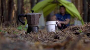 aromatisk kaffe är beredd i en gejser kaffe tillverkare. en man är vilar i en tält i de skog i de bakgrund. resa och friska livsstil begrepp video