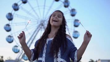 A beautiful girl with long hair holds fireworks in her hands and rejoices standing on the background of a ferris wheel. slow motion. video