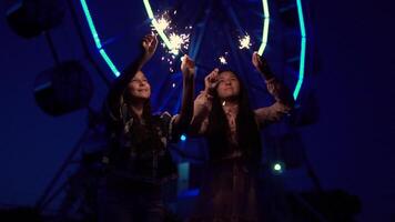 Two young girls with fireworks in their hands on the background of a ferris wheel. slow motion. video