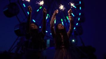 Due giovane ragazze con fuochi d'artificio nel loro mani su il sfondo di un' Ferris ruota. lento movimento. HD video